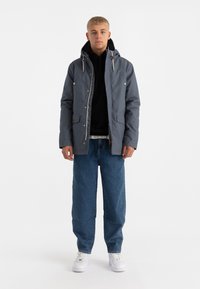 Giacca impermeabile grigia con cappuccio, chiusura a zip e due tasche frontali, indossata sopra una felpa nera con cappuccio e abbinata a jeans blu larghi e sneakers bianche.