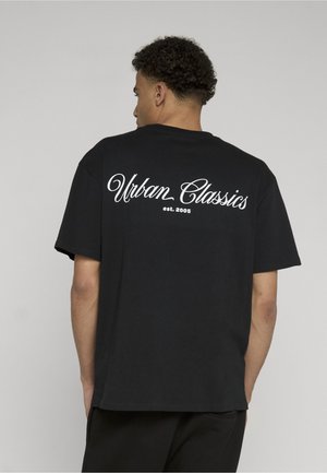 Jeune homme aux cheveux bouclés portant un t-shirt noir avec le texte blanc « Urban Classics est. 2005 » imprimé dans le dos.