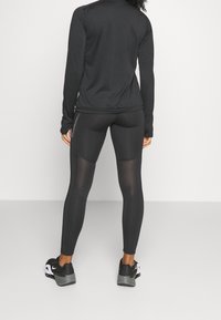 Černý dlouhý rukáv sportovní top s hladkou texturou. Černé legíny s průhlednými panely, doplněné černými sportovními botami s bílými akcenty.