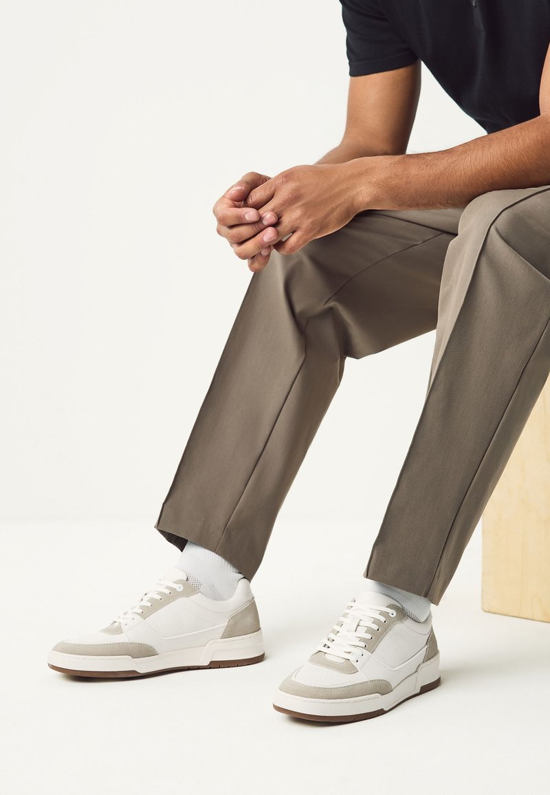 Personne assise portant un pantalon beige, des baskets blanches avec des accents gris clair et des chaussettes blanches, mains jointes reposant sur le genou.