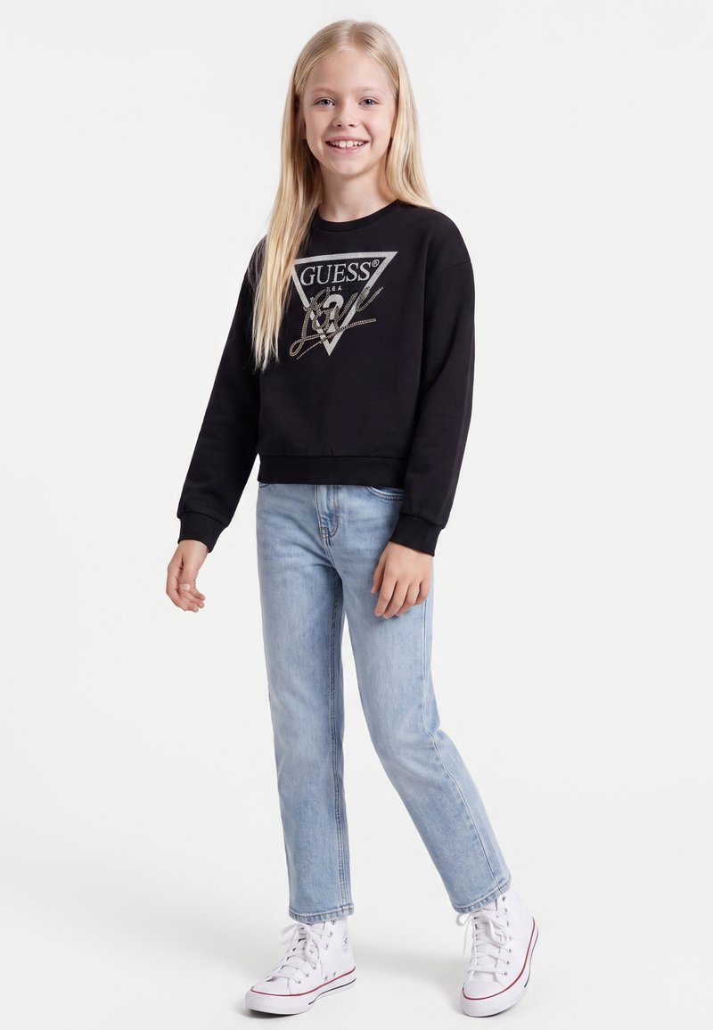 Fille souriante aux longs cheveux blonds portant un sweat-shirt noir Guess, un jean bleu clair et des baskets montantes blanches, debout sur un fond blanc.