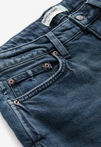 Jean en denim bleu foncé étiqueté "Relaxed Boyfriend", montrant la poche avant, la boucle de ceinture, le bouton et les rivets métalliques sur fond blanc.