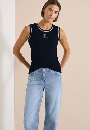 Femme portant un haut sans manches bleu marine avec le texte "ocean" et un jean taille haute à rayures bleu clair, debout les mains derrière le dos.
