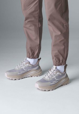 Person wearing beige and gray sneakers with white socks and brown cuffed pants standing on a light gray floor.