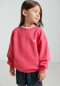 Różowy sweter z dzianiny z prążkowanymi mankietami i dekoltem, z białym logo "AMERICAN VINTAGE". Noszony z ciemnymi, teksturowanymi szortami.