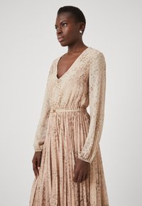 Φόρεμα με μακριά μανίκια και V-λαιμόκοψη από διαφανή ύφασμα με κωδικό λεοπάρ. Πλισαριστή φούστα, ελαστικοί καρποί και λεπτομέρεια με ζώνη σε χρυσό τόνο.