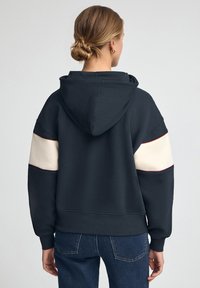 Mörk marinblå hoodtröja med en mjuk textur, med ljuskrämfärgade och vinröda detaljer på ärmarna. Avslappnad, oversized passform med huva.