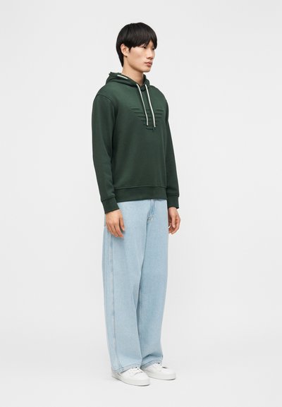 Άνδρας που φοράει σκούρο πράσινο φούτερ με ανάγλυφο σχέδιο, ανοιχτό μπλε τζιν με ευρύ μήκος και λευκά αθλητικά παπούτσια, στέκεται σε ουδέτερη στάση μπροστά σε ένα απλό φόντο.