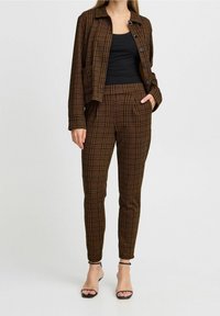 Veste à carreaux marron et pantalon assorti, avec une coupe décontractée, fermeture à bouton, et un haut noir, associés à des sandales à talons noires.