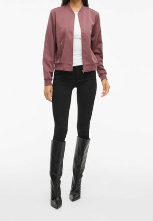 Frau trägt eine altrosa Bomberjacke, weißes Hemd, schwarze Skinny-Hosen und hohe schwarze Lederstiefel und steht vor einem weißen Hintergrund.