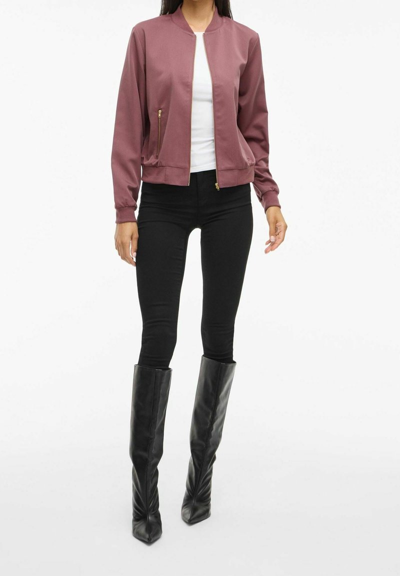 Femme portant une veste bomber mauve, une chemise blanche, un pantalon skinny noir et de grandes bottes en cuir noir, debout devant un fond blanc.