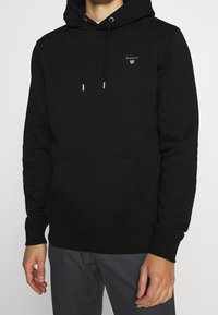 Zwarte katoenen hoodie met een voorvak, voorzien van een klein rood GANT-logo en zilveren oogjes voor het trekkoord.