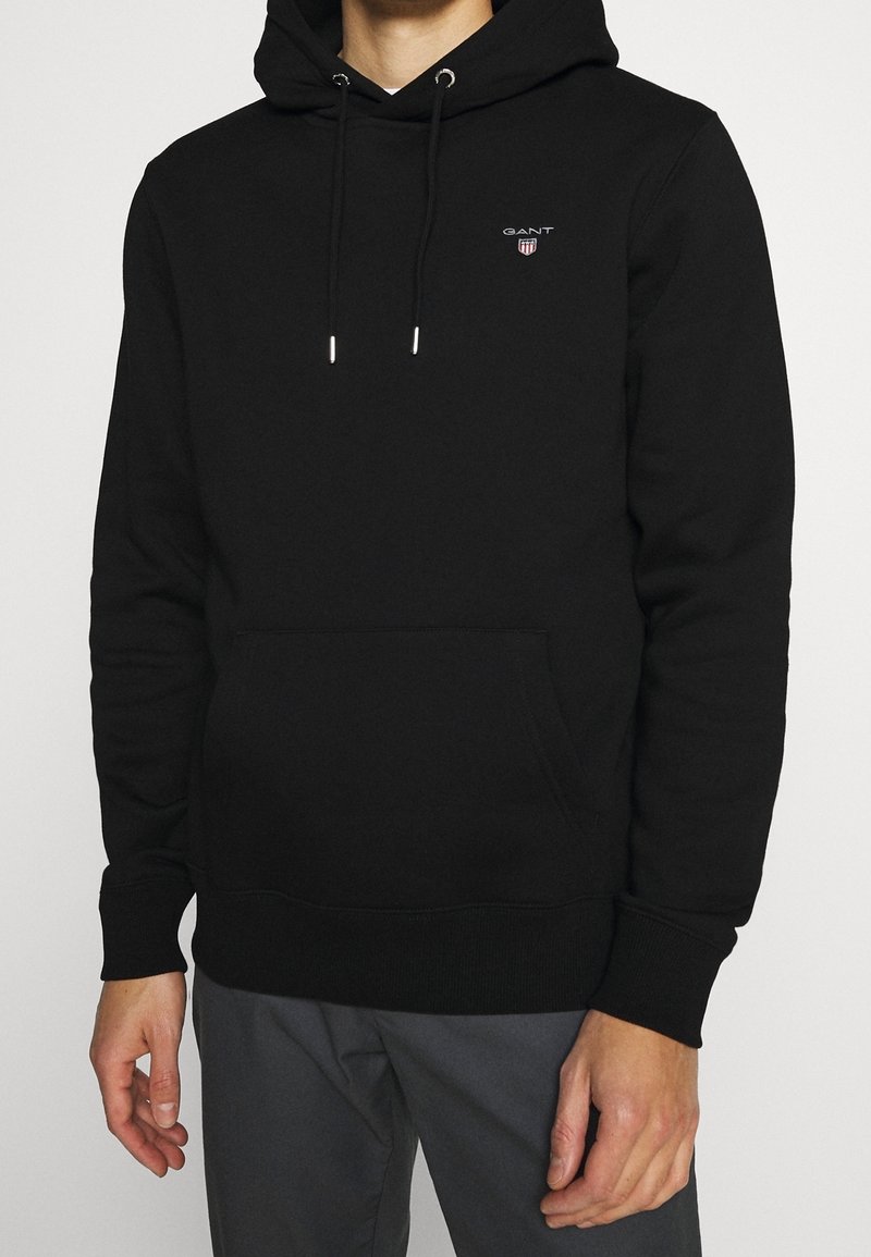 Zwarte katoenen hoodie met een voorvak, voorzien van een klein rood GANT-logo en zilveren oogjes voor het trekkoord.