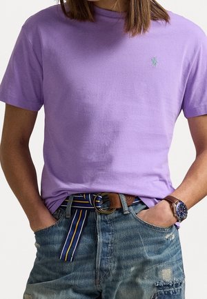 Personne portant un t-shirt lavande à col rond, une ceinture bleue à rayures, un short en jean déchiré et une montre-bracelet avec un bracelet en cuir marron.