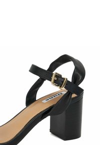 Sandale noire à talons hauts avec une sangle courbée, une boucle en métal doré, une texture en cuir lisse et un talon épais.