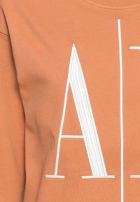 Tričko z měkké terakotové látky s velkým bílým vyšívaným písmenem "A" a svislými bílými pruhy na přední straně.