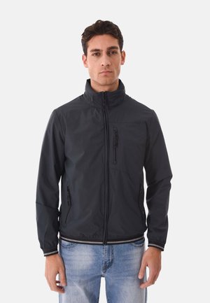 Uomo che indossa una giacca nera con cerniera, dotata di tasche con zip sul petto e sui lati, in piedi davanti a uno sfondo chiaro uniforme.