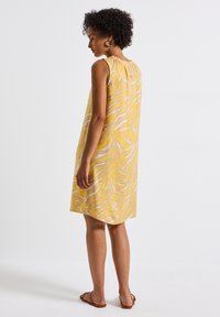 Gelbes ärmelloses Kleid mit einem weißen Zickzackmuster, knielang, lockere Passform und Raffung am Ausschnitt. Sandalen sind sichtbar.