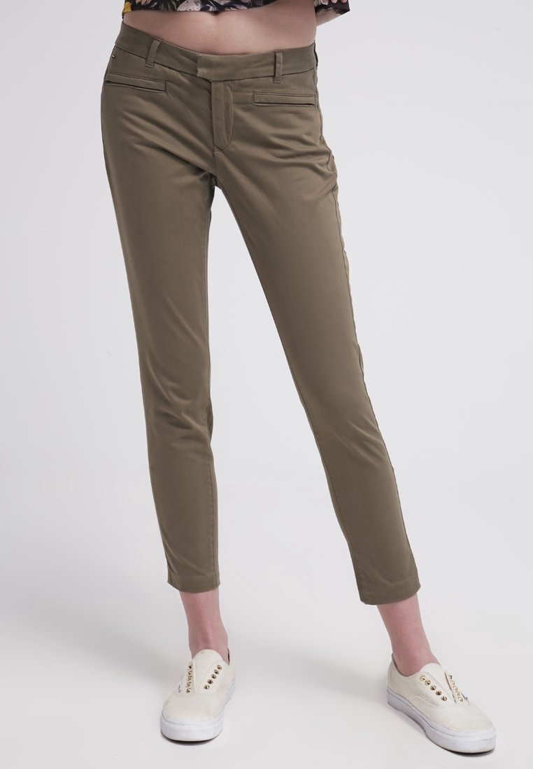 Olive green slim-fit pants with a zip fly, two front pockets, and a smooth texture. Paired with white sneakers featuring round studs.