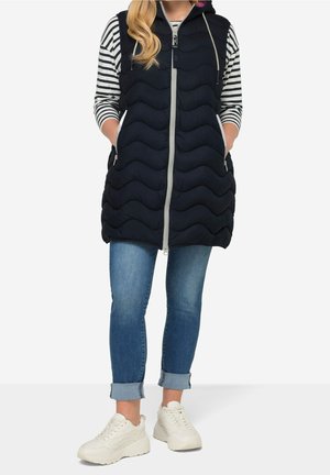 Kvinde iført en sort quiltet ærmeløs hættevest over en stribet langærmet skjorte, opkrogede blå jeans og hvide sneakers.