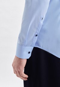 Camicia azzurro chiaro con polsini abbottonati, caratterizzata da una tessitura morbida e un sottile accento ricamato sul polsino.