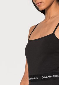 Svart linne med tunna axelband, tillverkat av mjuk bomull, har ett märkesband i vitt med texten "Calvin Klein Jeans".