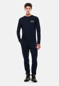Maglietta a maniche lunghe color navy con logo EA7, abbinata a pantaloni sportivi navy con strisce laterali. Il modello indossa scarpe da ginnastica nere.
