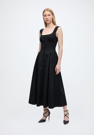 Un mannequin se tient devant un fond uni, vêtu d’une robe midi noire sans manches, boutonnée sur le devant, et de chaussures à talons hauts à brides.