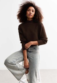 Pullover in maglia a coste marrone con colletto alto, abbinato a jeans a gamba larga in denim chiaro e sneaker bianche, presentato su uno sfondo semplice.