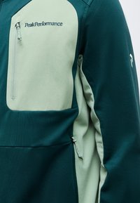 Δίχρωμο πράσινο μπουφάν Peak Performance με φερμουάρ στην τσέπη του στήθους και λογότυπο στο πάνω αριστερό μέρος του στήθους και στο μανίκι.