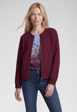 Blonde woman wearing a maroon zip-up jacket over a graphic blue and purple t-shirt with dark blue jeans against a plain background.