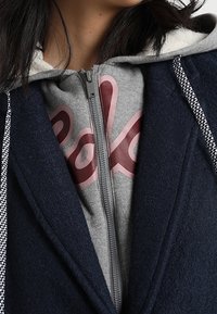 Námořnický kabát z vlněné směsi se zipem vpředu, pod kterým je šedý svetr s červeným logem. Doplňky zahrnují kapuci a vzorované šňůrky.