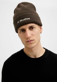 Bruine geribbelde beanie met witte tekst "JJ Studios", gedragen met een zwarte gestructureerde trui. De muts zit strak, waardoor het gebreide patroon goed zichtbaar is.