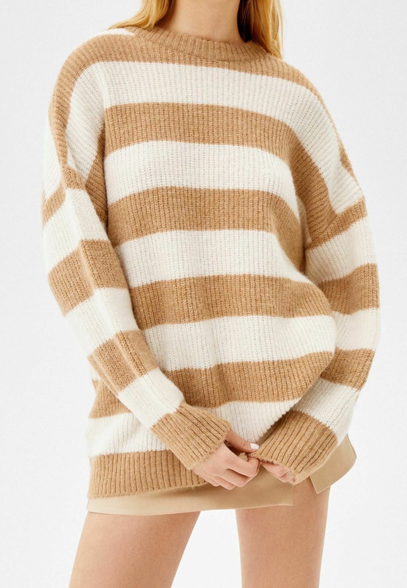 Personne portant un pull en maille oversize beige et blanc à rayures horizontales, manches longues et poignets côtelés, associé à une jupe beige.