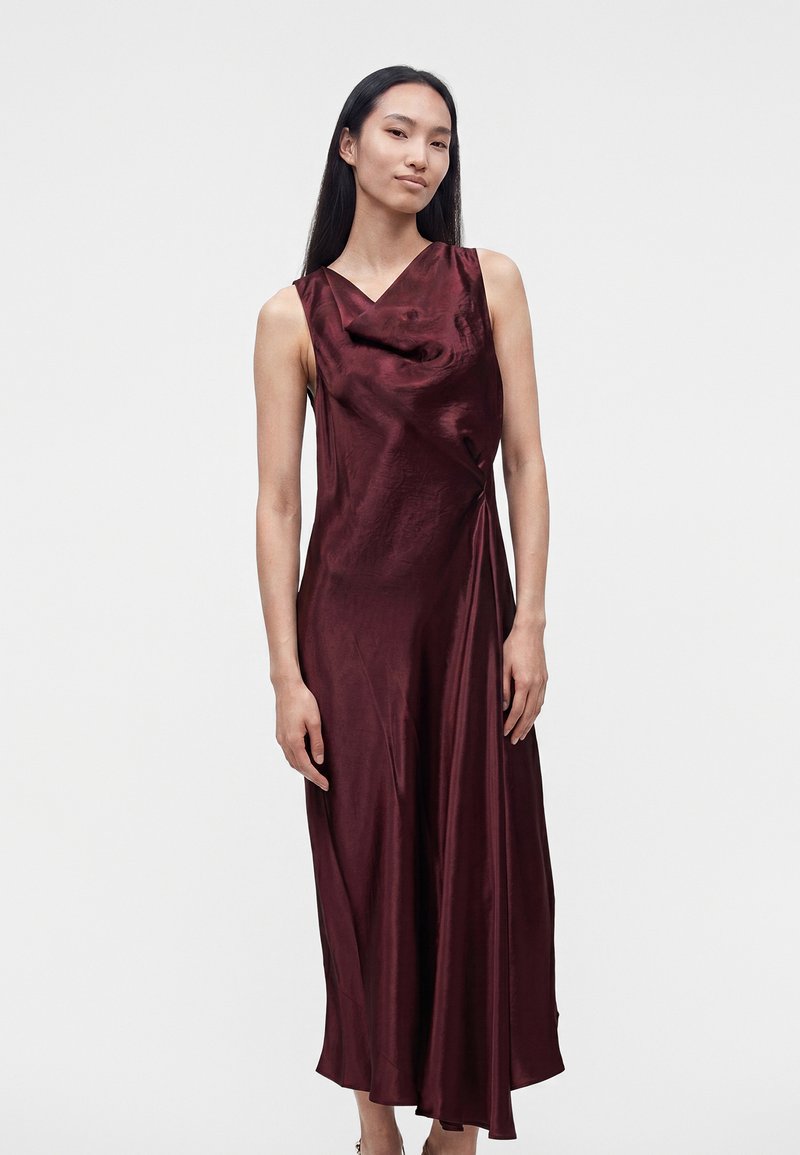 Femme debout dans une robe longue sans manches en satin rouge foncé avec un col drapé, de face devant un fond uni.