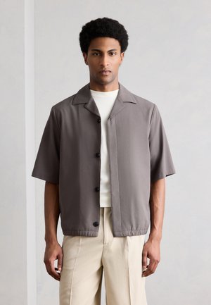 Moški s kratkimi kodrastimi lasmi nosi taupe srajco s kratkimi rokavi s gumbi preko bele majice in bež hlače, stoji ob svetli enobarvni steni.