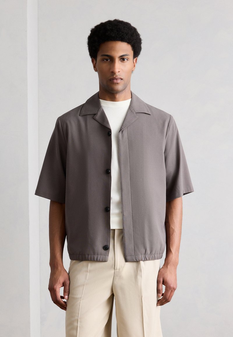 Άνδρας με κοντά, σγουρά μαλλιά φοράει ταμπά κοντομάνικο πουκάμισο με κουμπιά πάνω από λευκό μπλουζάκι και μπεζ παντελόνι, όρθιος μπροστά σε απλή, φωτεινή τοίχο.