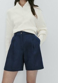 Pull en tricot crème avec un col montant, associé à un short bleu marine ajusté avec un bouton à l'avant et des poches latérales.