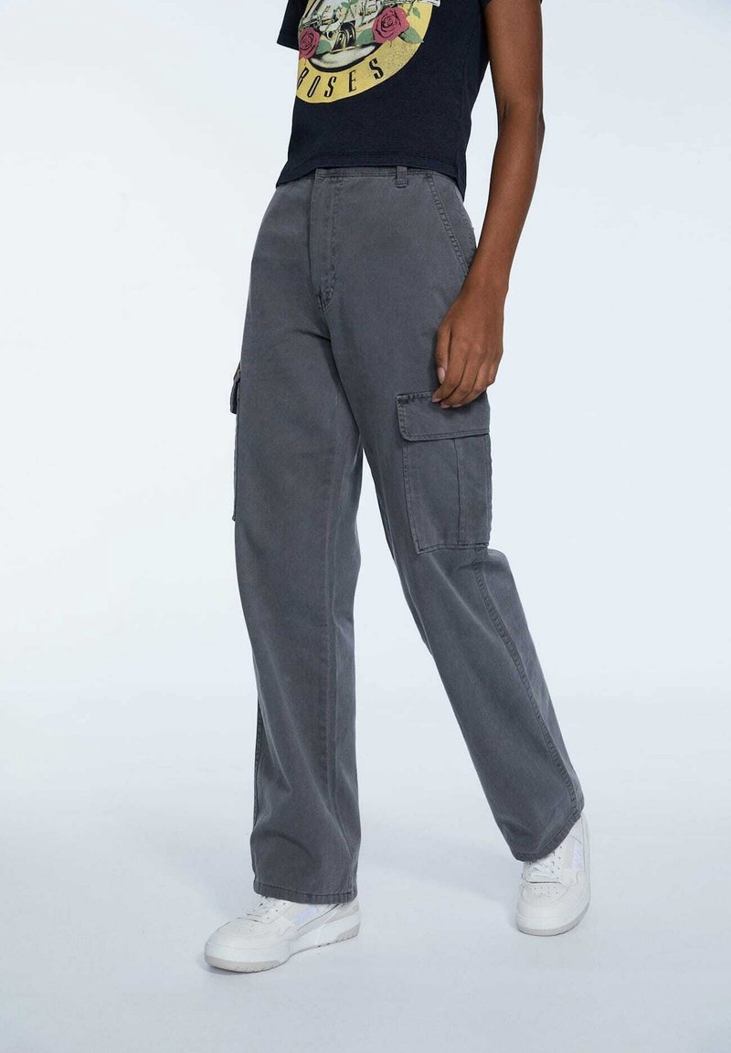 Personne portant un pantalon cargo gris, des baskets blanches et un t-shirt noir avec un motif jaune et rouge.
