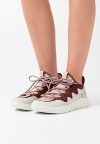 Nízké tenisky s burgundským semišovým svrškem, béžovými akcenty, texturovanou bílou podrážkou a kontrastními šněrovadly se žlutými špičkami.