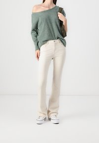 Lichtgroene oversized breiende trui met één schouder bloot, gecombineerd met crèmekleurige rechte jeans en witte sneakers, staand op een effen achtergrond.