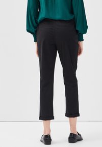 Pantalon noir court avec une texture lisse, doté de revers roulés. Porté avec un haut vert à manches longues et des chaussures noires à finition texturée.
