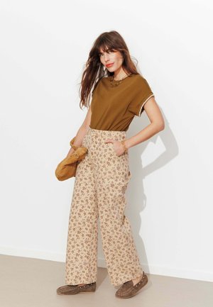 Femme aux longs cheveux bruns portant un haut marron, un pantalon large à motif floral, des mocassins marron et tenant un sac à main marron.