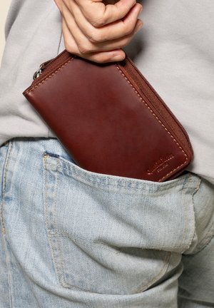Rjavo usnjeno denarnico z zadrgo, pravokotne oblike in diskretno šivane detajle, delno pospravljeno v svetlo modri džins žep.