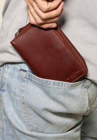 Brown leather wallet with a zip closure, rectangular shape, and subtle stitching details, partially tucked into light blue denim pocket.