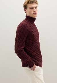 Bordeauxroter Pullover mit Zopfmuster, hohem Kragen, strukturierter Oberfläche, gerippten Bündchen und Saum. Über hellen Hosen getragen.