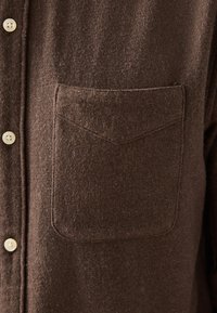Chemise en laine marron avec une poche poitrine frontale unique et des boutons de couleur crème le long du côté gauche.