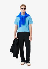 Camisetta azzurra a maniche corte, maglione blu annodato intorno al collo, pantaloni neri, che tiene una giacca nera, indossa occhiali da sole e scarpe chiare.