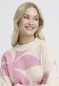 Pull beige clair avec grand motif floral rose, finition texturée, encolure ronde et poignets côtelés.