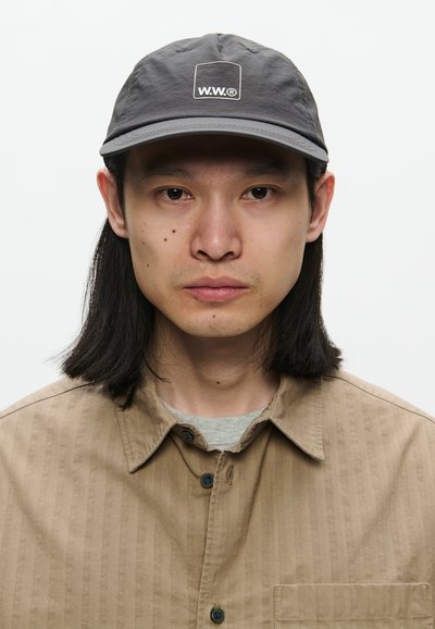 Azijski muškarac s kestenjastom dugom kosom nosi sivi šešir s logom "W.W.®" i svijetlosmeđu košulju preko sive majice, okrenut prema naprijed.